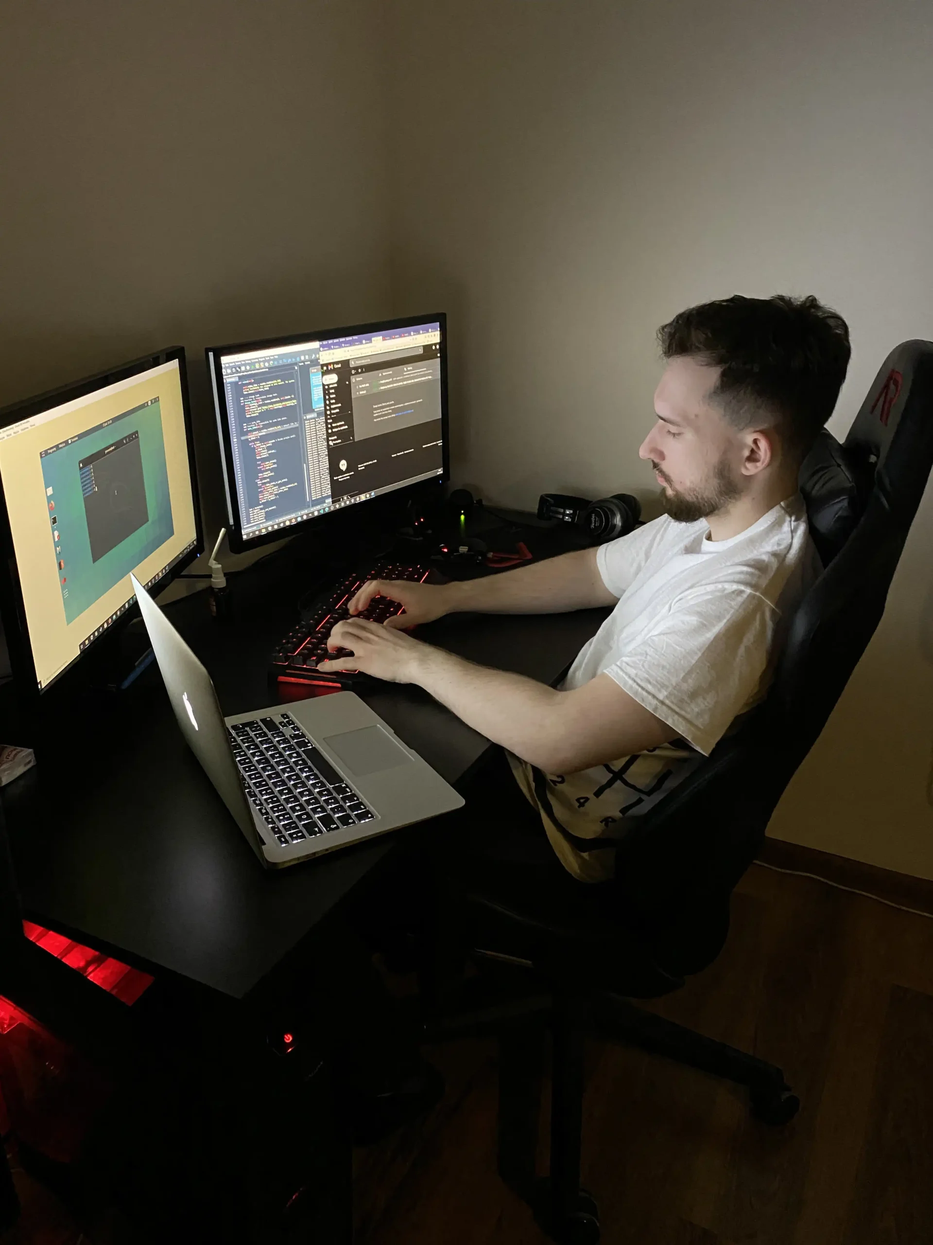 Osoba pracująca przy biurku z trzema monitorami, laptopem i podświetlaną klawiaturą; na ekranach widać środowisko techniczne oraz kod.